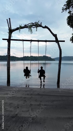 Aerial drone shot of a romantic couple in love sitting together on rope swing at sunset beach, young man and woman on holidays or honeymoon.