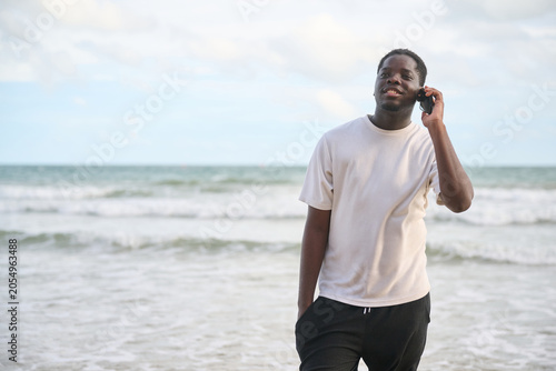 Young man in a white t-shirt and black pants stands on a beach, talking on his phone. The ocean and sky are visible in the background