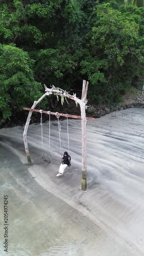 Moslem asian woman person enjoys a relaxing moment on a wooden swing at the beach in Weh island Sabang Indonesia, overlooking the ocean and the horizon on a sunny day.  Travel at the resort