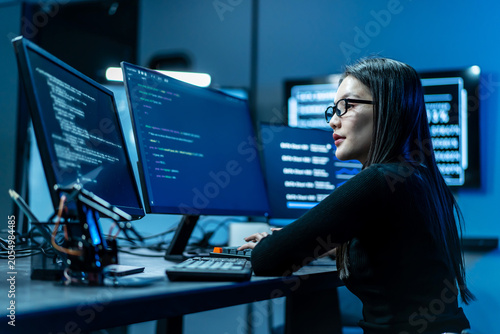 Foto Focused young Asian female software engineer writing code on multiple monitors in a dark modern IT office