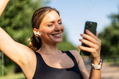 Young sporty woman wearing headphones, enjoying music on her phone, dancing and smiling during an outdoor workout in the public park, radiating energy and happiness.
