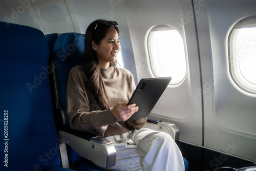 Young woman sitting by the airplane window and using a tablet during a flight, smiling in a comfortable seat. Ideal for travel, business trip, remote work, and modern lifestyle themes.