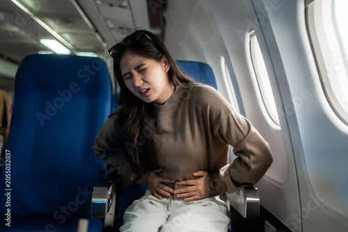 Young woman sitting by an airplane window looks unwell and holds her stomach, showing travel sickness or abdominal pain during a flight.