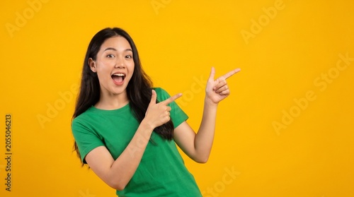 A young woman in a green t-shirt poses against a bright yellow background, smiling with an open mouth and pointing to the right with both index fingers.