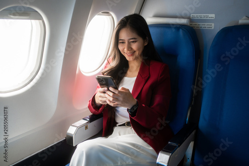 Young woman in a red blazer sits by an airplane window, talking on her phone during a flight. calm travel scene with natural light, business trip, communication, and modern air travel.