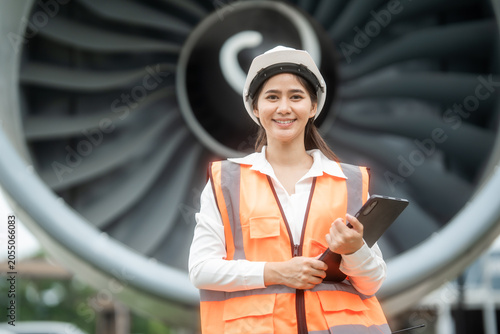 Smiling female engineer in safety vest and helmet holding a tablet in front of a large jet engine at an airport, showing aviation work, inspection, maintenance and travel industry.