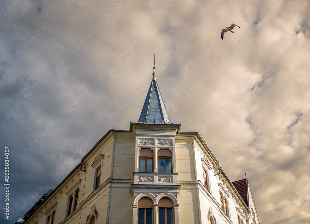 Obraz premium Historic corner building with soaring spire and flying bird against textured clouds creating timeless European street photography composition
