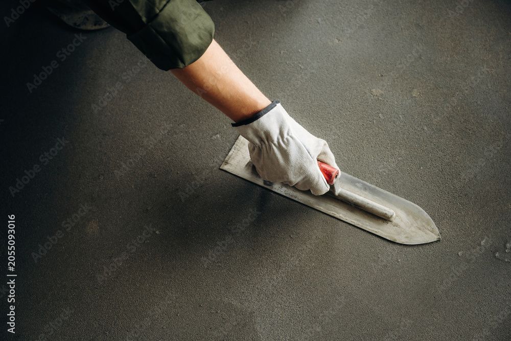 Naklejka premium Hand of a construction worker wearing a glove using a trowel to smooth wet concrete surface on a floor during renovation or construction project
