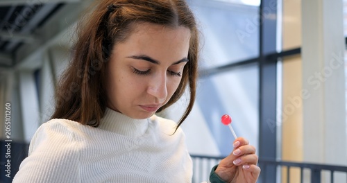 Konstfotografi Close portrait of young woman near window holding lollipop with thoughtful expression, soft daylight illuminating face and textured sweater, intimate quiet mood in transit corridor