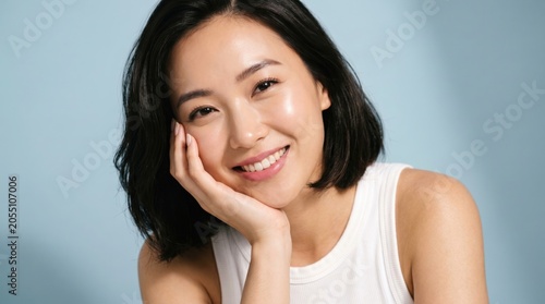 Portrait of a beautiful young Asian woman with glowing clear skin and a radiant smile, leaning on her hand against a blue background, representing skincare, wellness, and natural beauty.