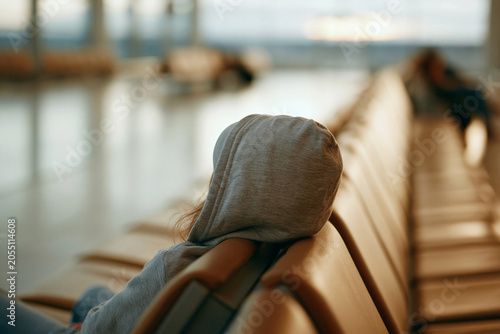 Child sits quietly, hooded, on airport bench. Soft focus shows distant travelers, quiet hum of travel. Sunlight glows, warm, low, on waiting skin and seat. Emotion is stillness, comfort