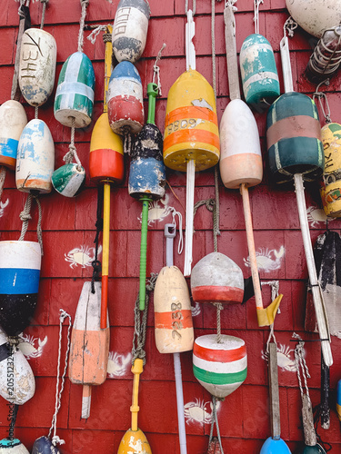 Cuadro en lienzo Collection of colorful, weathered lobster buoys hanging on a bright red fishing shack wall