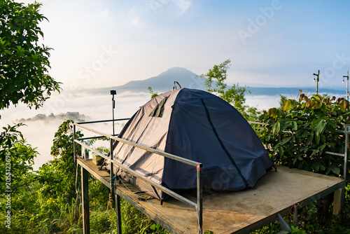 Mountain Campsite Sunrise: A cozy tent set up on an elevated platform offers a breathtaking view of a misty mountain valley at sunrise, surrounded by lush green foliage.