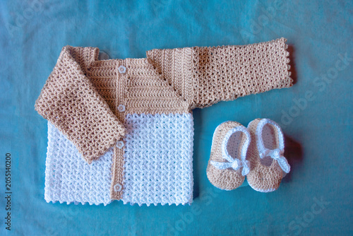 Crocheted baby jacket and booties in beige and white on a blue background, symbolizing warmth and handmade care