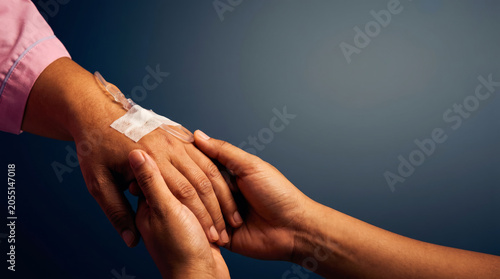 Hands holding patient hand with an IV line attached on dark blue background.