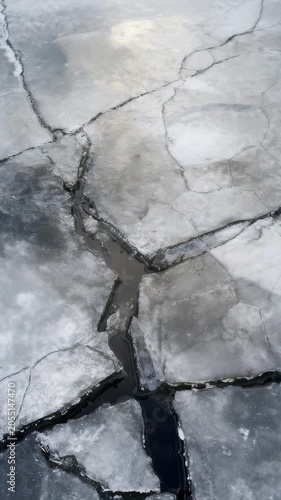 Cracked frozen lake surface with visible dark water lines, showcasing dramatic winter textures and natural ice patterns. Abstract nature background for climate change themes