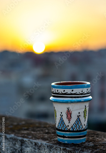 Traditional Moroccan ceramic tea glass on a rooftop ledge with a blurry sunset over the Meknes skyline, Morocco