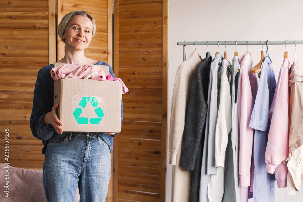 Fototapeta premium Woman standing with a cardboard box full of clothes for recycling. Sustainability concept for garment donation. Eco friendly lifestyle choices, mindful consumption and fashion waste reduction.