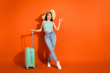 Young stylish female traveler posed against a vibrant orange background with suitcase ready for an exciting vacation
