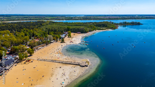 Wide aerial drone view of a sandy lake beach with a wooden pier, vibrant blue water, surrounded by lush green forest and resort buildings on a bright sunny summer day.