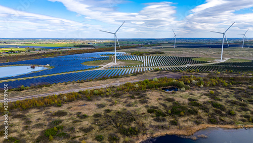 Wide aerial drone view of a modern solar farm with rows of blue photovoltaic panels and tall wind turbines scattered across green fields under a blue sky with distinctive clouds.