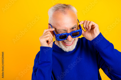 Playful senior man winking and smiling in blue sweater and sunglasses against a bright yellow background