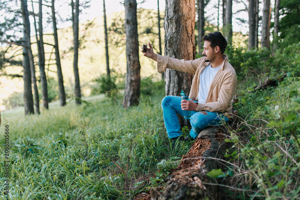 Obraz premium A hiker takes selfies while resting on a fallen wooden log, breathing fresh air and enjoying the colorful, sunny forest.