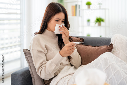 A woman holding fever thermometer suffering from a cold, the flu, or allergies is blowing her nose with a tissue while lying on a sofa in the living room.