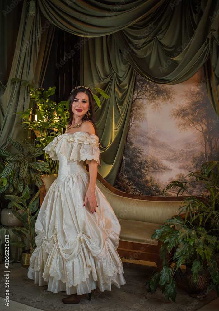 Fototapeta premium Elegant mature woman in a white lace off the shoulder dress posing in a classic interior, age female model wearing a fashionable evening gown, vintage style fashion concept