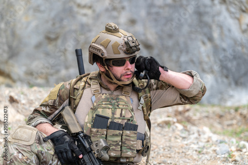 Military soldier in camouflage uniform using radio communication device during tactical mission outdoor. Tactical army officer listening to message on transceiver while hold gun during combat training