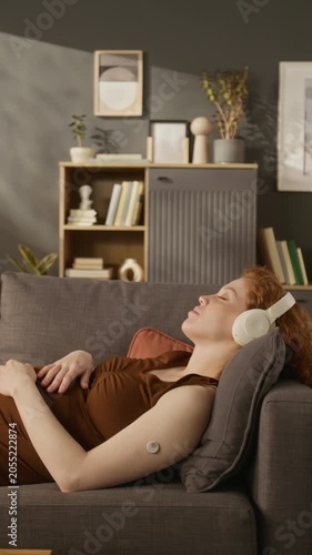 Vertical shot of pregnant Caucasian young adult woman with glucose monitor lying on sofa and listening to music. Perfect for marketing campaigns on gestational diabetes and prenatal care