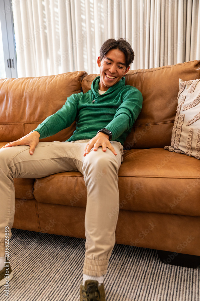 Fototapeta premium Asian man sitting on leather couch in living room, smiling, wearing green pullover and smartwatch