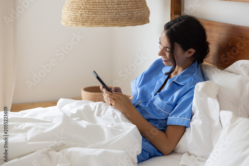 Woman relaxing and smiling on bed with white pillows and wooden headboard, holding smartphone