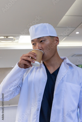 Adult Korean man wearing white lab coat and surgical cap drinking from paper cup in clinic