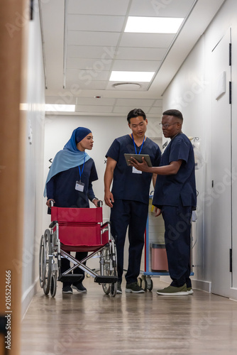 Diverse medical team checking tablet beside red wheelchair in clinic hall, wearing navy scrubs IDs