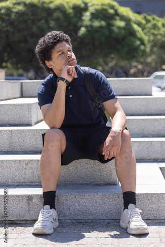 Man sitting on steps in plaza resting chin wearing navy polo, backpack strap, wrist device
