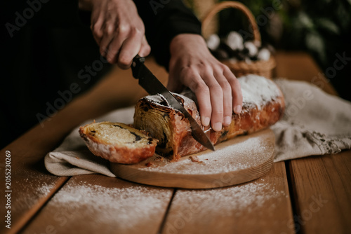Close-up of baker hands slicing a fresh homemade sweet pastry roll with a knife on a wooden board, rustic food styling.
