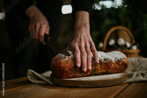 Close-up of baker hands slicing a fresh homemade sweet pastry roll with a knife on a wooden board, rustic food styling.