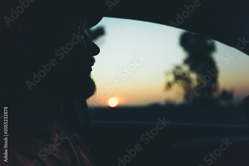 Bild auf Leinwand Silhouette of bearded man looking through car window at golden sunset during a p