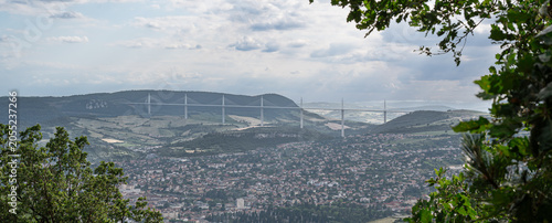 Millau Viaduct rising above the town of Millau in Aveyron, France. Iconic cable stayed bridge spanning the Tarn Valley framed by woodland and dramatic summer clouds.