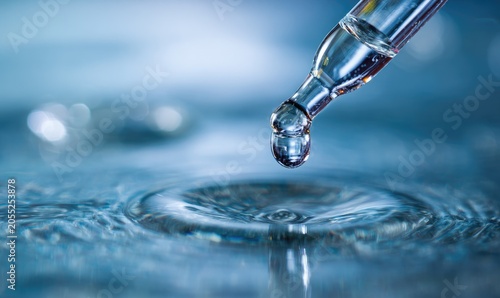 Close-up of a glass dropper with a single drop of liquid falling, creating ripples on the water's surface, reflecting light