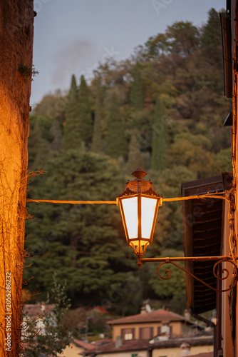 An old-fashioned, ornate street lamp glows warmly, suspended between two ancient stone walls, casting a romantic ambiance over a distant hillside village as dusk settles