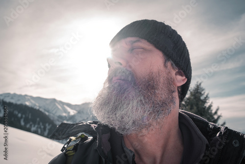 Close-up portrait of a bearded mature man looking up outdoors against snow-covered mountains and sun. Commercial stock image for travel, lifestyle, winter adventure, and outdoor recreation concepts.