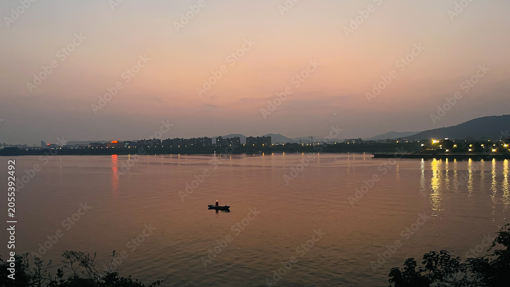 Fototapeta premium Tranquil twilight silhouette of a small boat on a vast river with a distant illuminated city skyline reflecting on the water