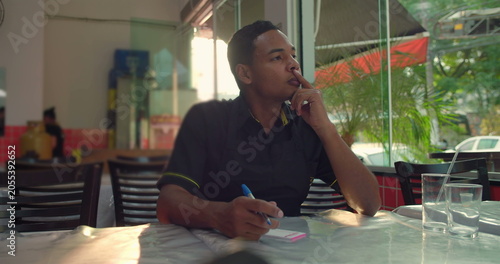 Fotografie Latin waiter of African descent sits at restaurant table thoughtfully holding pe