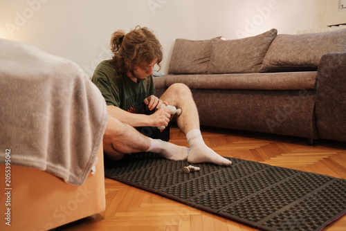 Young man using a massage gun on leg muscles after training. Concept of sports recovery, muscle soreness relief, physiotherapy, home workout and healthy habits.