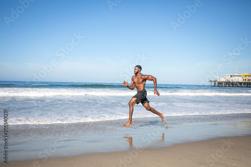 Man running on a sunny beach near the ocean, pier in the background.