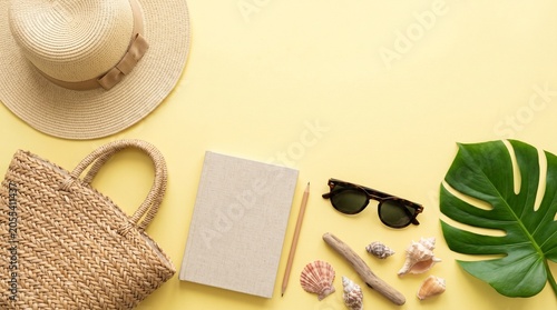 Summer travel flat lay with notebook, pencil, straw hat, woven bag, sunglasses, and sea shells on yellow background. Vacation planning concept.