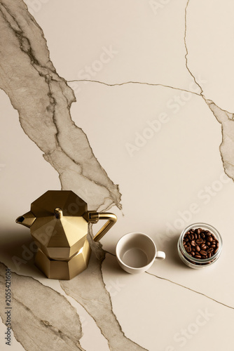 Overhead view of gold moka pot beside espresso cup and glass jar of roasted coffee beans on cream marble surface.