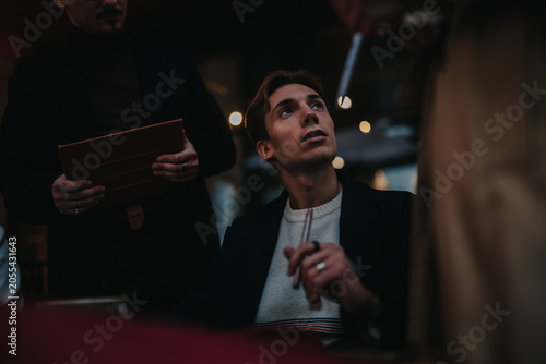 Slika na platnu A young man holding a pen looks up while speaking with staff in a dim restaurant